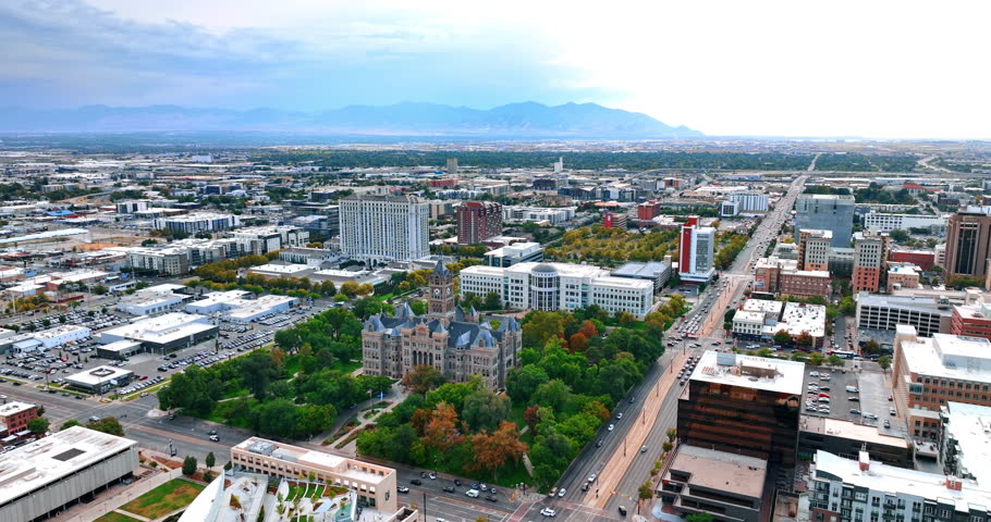 Flying closer to the beautiful building of the Salt Lake City and County Building surrounded by the lovely park. Vast urban landscape with mountain silhouette at backdrop.