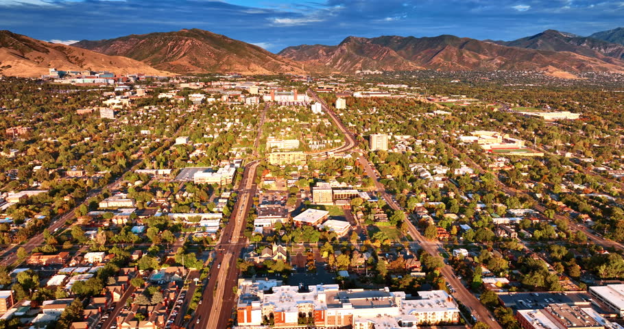Bright sun of sunset dazzling the dense urban landscape of Salt Lake City, Utah, USA. Drone flight over the green neighborhoods approaching the bare mountains.