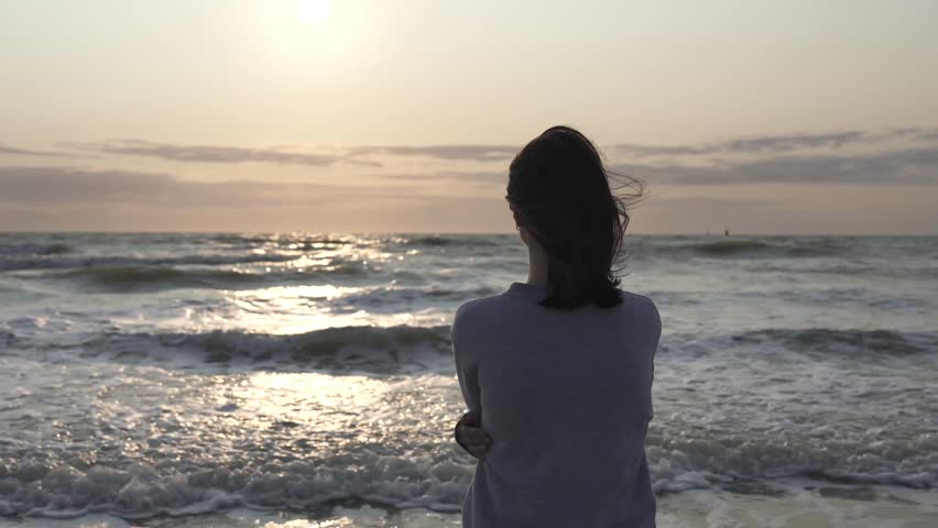 Back view of a lonely girl standing in the water and watching stormy sea waves.
