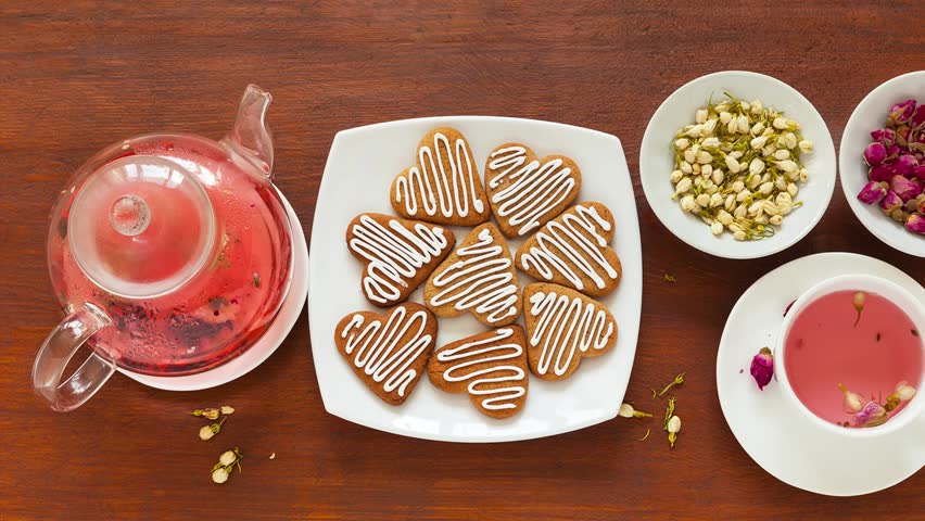 Video of freshly brewed floral tea with jasmine flowers and rosebuds in glass teapot and white cup on wooden table. Handcrafted homemade heart-shaped cookies are nearby. Top view, close-up, flat lay