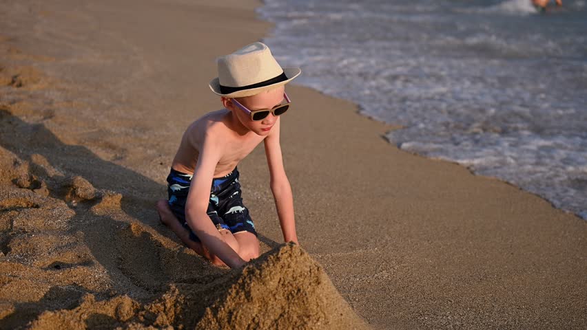 Boy builds sand castle on sandy beach at tropical resort, watching incoming waves. Family vacation at seaside, summer holidays