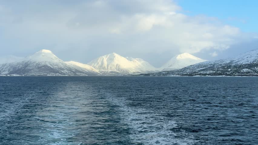 A boat makes its way through a fjord in Norway, leaving a wake in the clear water. Snow-covered mountains are visible along the shore under a blue sky.