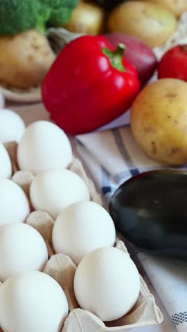 Fresh eggs on a checkered cloth and a mesh bag overflowing with broccoli, potatoes, tomatoes, and other vegetables on a dark surface.