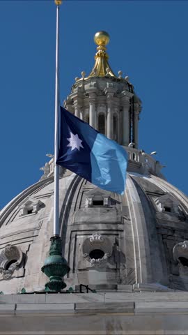 Flag is waving atop the State Capitol building in St Paul, Minnesota, showing the state symbol under a blue sky.