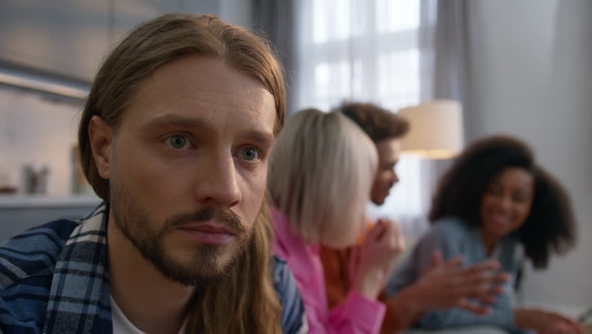 Stressed sad Caucasian man sitting couch home thinking about problems depressed male feeling separated ignoring friends while multiracial African women men talk together friendship loneliness anxiety
