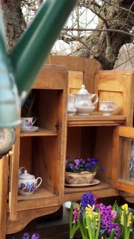 A close-up of a wooden cupboard standing in a blooming spring garden, filled with beautiful ceramic plates and pots of daisies. The soft sunlight highlights the textures of wood and clay against the vibrant green background.
