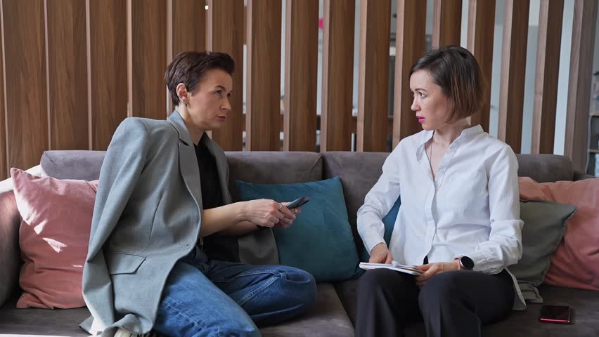Female business colleagues discussing and working with paperwork while seated on couch in stylish office. Colleagues, paperwork, couch, office, discussion.
