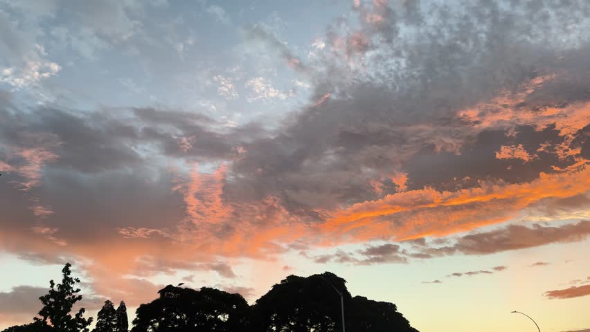 Vibrant sunset sky with dramatic orange and blue clouds over a suburban landscape, featuring silhouettes of trees and buildings at dusk.