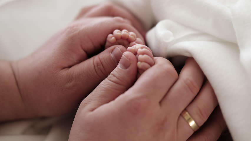 Father Hand Supports Tiny Feet Of Newborn Baby. Soft Blanket Surrounds Feet.