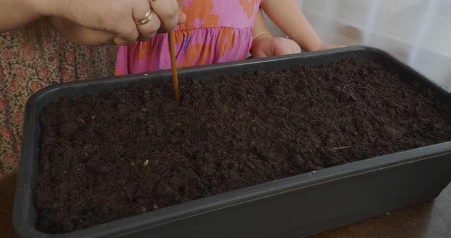 Mother and daughter planting seeds together, using a small wooden stick to make holes in the rich soil inside a planter box, representing an educational family activity and urban gardening.