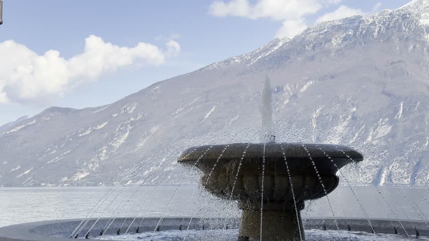A stone fountain sprays water on the lakeside promenade at Lake Garda, Italy. Water droplets cascade down the sculpted stone basin in a close-up view. In the background, the deep blue lake and dramatic snow-capped Alpine mountains are visible under a partly cloudy blue sky. Shot in 4K HDR on a bright sunny day. Perfect for nature, travel, water, and Italian landscape projects.