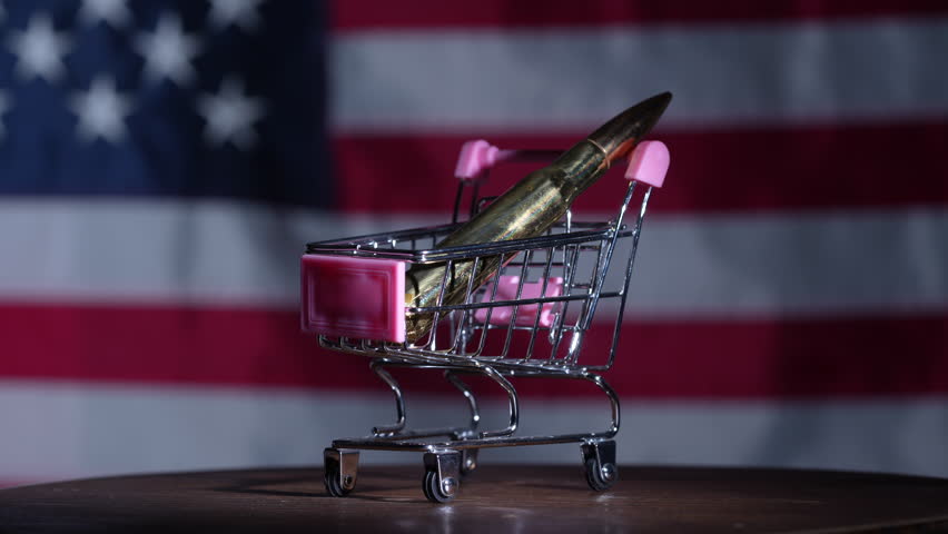 Close up of a bullet placed in a miniature shopping cart against USA flag background symbolizing gun trade consumerism and political debate concept, rotates