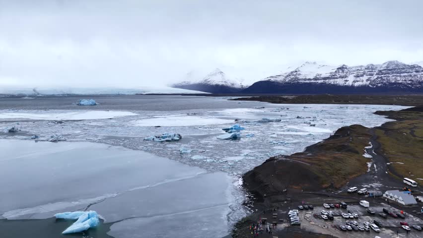 Aerial 4K video of Jökulsárlón showing floating icebergs in the lagoon, with tourist car park in view, surrounded by snowy landscape and glacial scenery in Iceland.