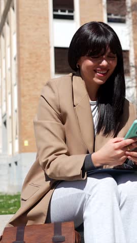 Happy female university student sitting on a bench outdoors, taking a selfie with her smartphone, smiling and showing a peace sign, enjoying a sunny day on campus between classes