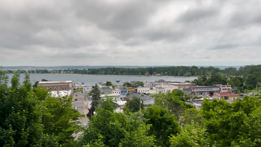 Aerial View of Harbor Springs, Michigan Waterfront from the Bluff. A steady high-angle shot overlooking the town of Harbor Springs, Michigan, from a hillside vantage point. The footage shows the lush green bluff leading down to the historic downtown, marina, and the tranquil harbor of Little Traverse Bay on a moody, overcast day.