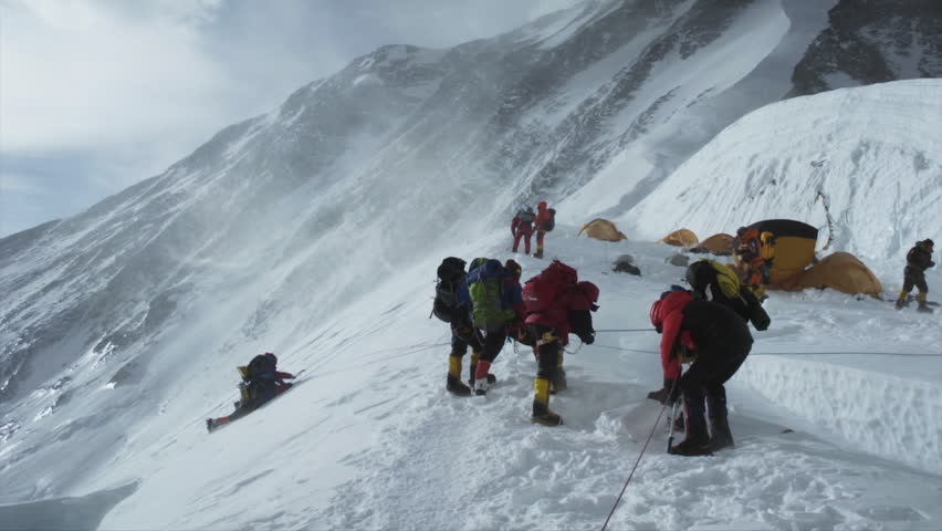 Group of alpinists roped together climbing a steep, snowy mountain path near a high altitude camp