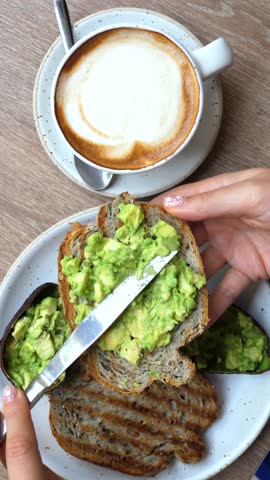 Top down view of a woman's hands using a knife to spread fresh mashed avocado on a slice of grilled rye toast, preparing a healthy and delicious breakfast served with a cup of hot coffee