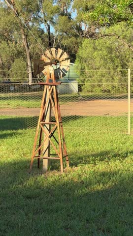 A decorative wooden windmill stands in a grassy backyard in rural Australia, with eucalyptus trees and a wire fence visible in the background. The rustic garden ornament adds country charm to the peaceful pastoral setting.