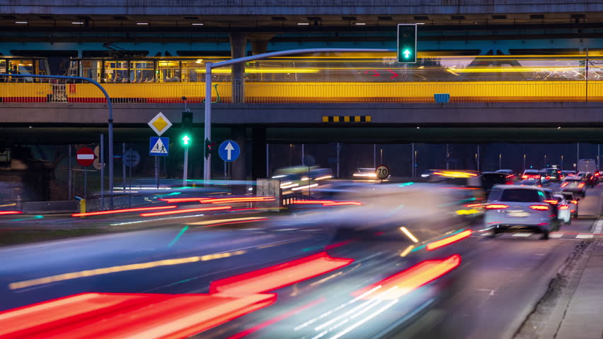 Timelapse of busy city traffic at intersection during evening. Cars move through roadway while others wait at red lights, and yellow trams pass across the bridge above. Multi-level urban transport scene with dynamic motion and glowing light trails
