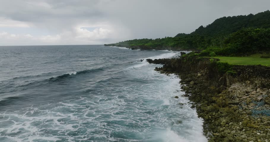Rocky shore with ocean waves crashing against the coast under cloudy sky. Siargao, Philippines.