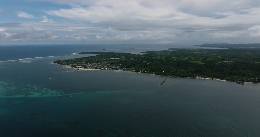 A coastal village lies along the shoreline with coral reef waters and cloudy sky over the sea. Siargao, Philippines.