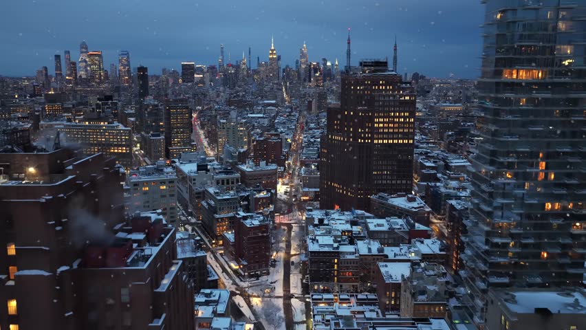 Aerial night view of New York City during snowfall, with illuminated streets, glowing office towers and calm winter traffic stretching through downtown skyline of manhattan. Slow drone flyover.