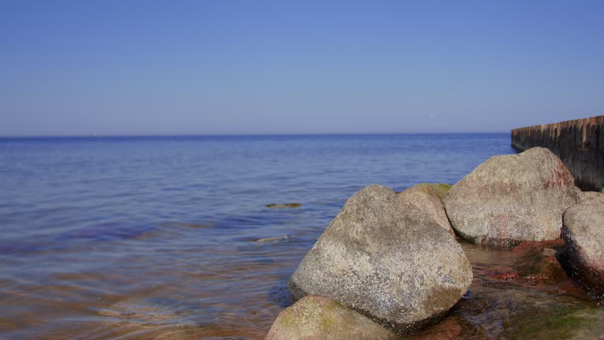 Sea waves washing over stony beach with wooden pier. Coastal landscape with rocky shore, water movement and structure. Summer seascape scene by seaside nature.