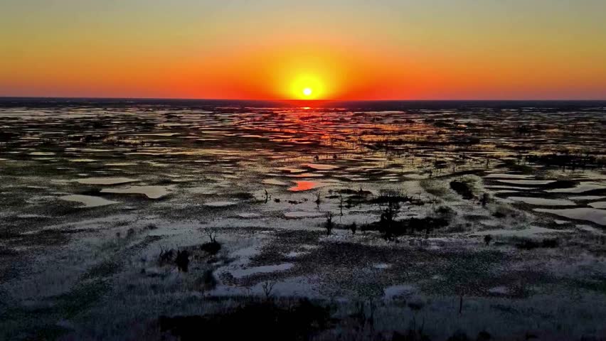 "Golden sunset over the Okavango Delta, featuring vast wetlands, reflective water channels, and a vibrant orange horizon