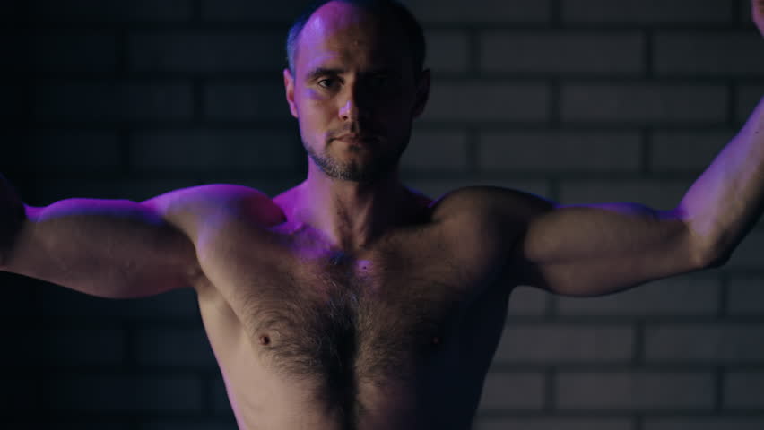 Athletic shirtless man tensing his arm muscles in a dark gym with dramatic lighting. The strong bodybuilder showing his powerful physique, showcasing strength, masculinity, and dedication
