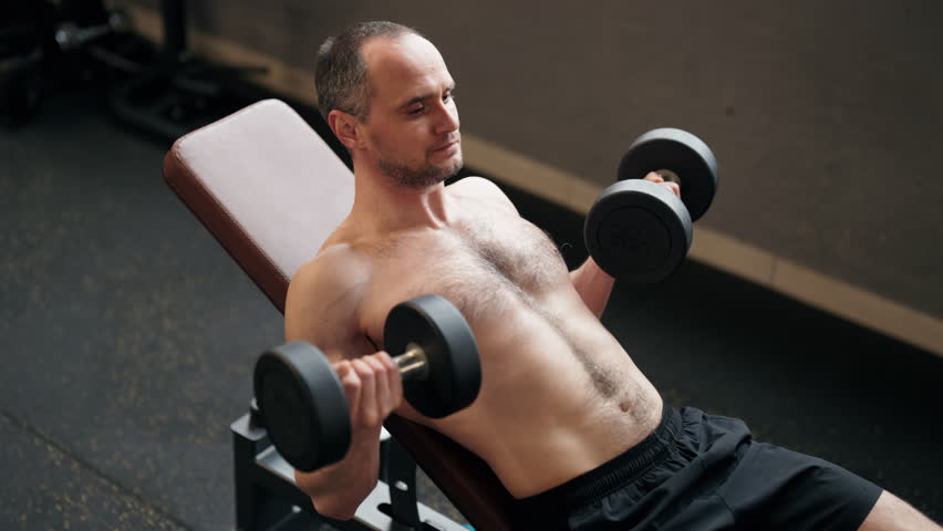 Strong athletic man with a bare chest lifting dumbbells while sitting on an incline bench during a strength training session, building his biceps and focusing on muscle growth in a gym