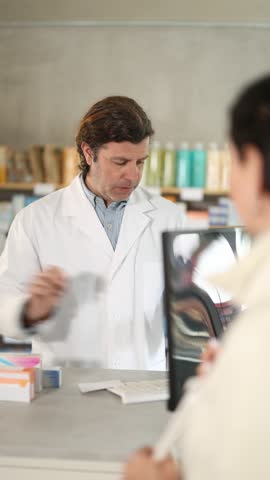 Male pharmacist stands behind the counter near the computer and communicates with a customer. Employee of a European pharmacy consults a woman and offers her pills according to her prescription. High