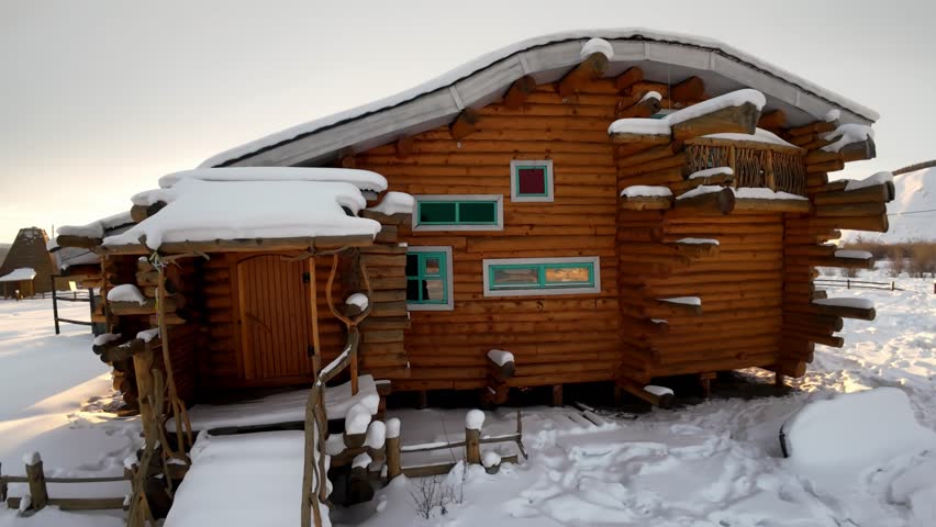 A rustic wooden cabin sits in a snowy landscape with mountains in the background as the sun sets.