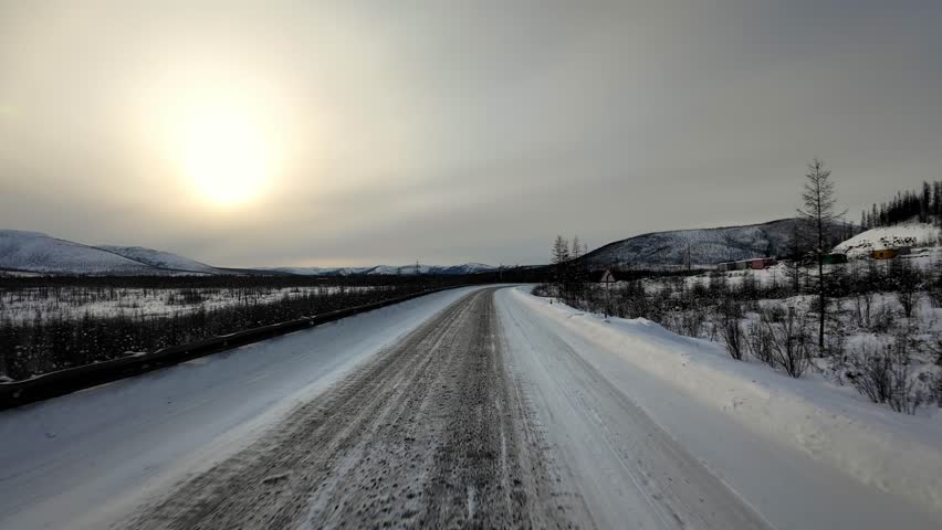A snow-covered road winds through a winter landscape with trees and mountains visible, captured during daytime.