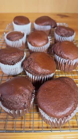 Vertical video - Closeup of a hand picking up one from the back of a homemade batch of freshly baked, soft, rich, chocolate brown muffins in paper cases, on a cooling grid.
