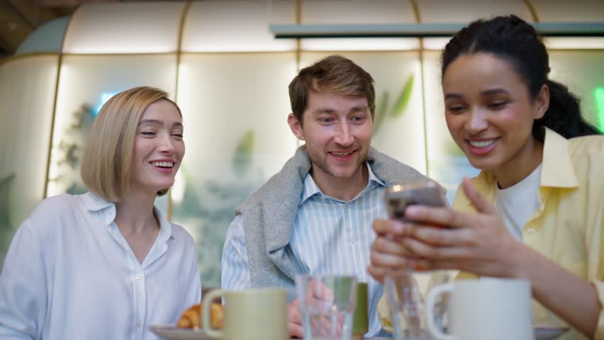 Three friends smiling and watching smartphone during cafe breakfast. Adult man and women sitting at table with coffee and pastry. Looking at screen together and enjoying moment. Friendship.