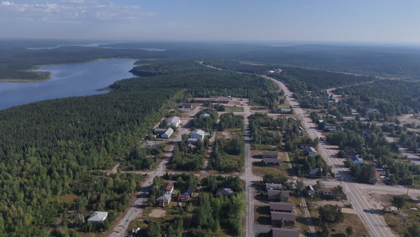 High aerial drone flyover of Lynn Lake Manitoba showing the highway leaving town, houses (some abandoned), surrounding boreal forest, and a lake on a summer day.