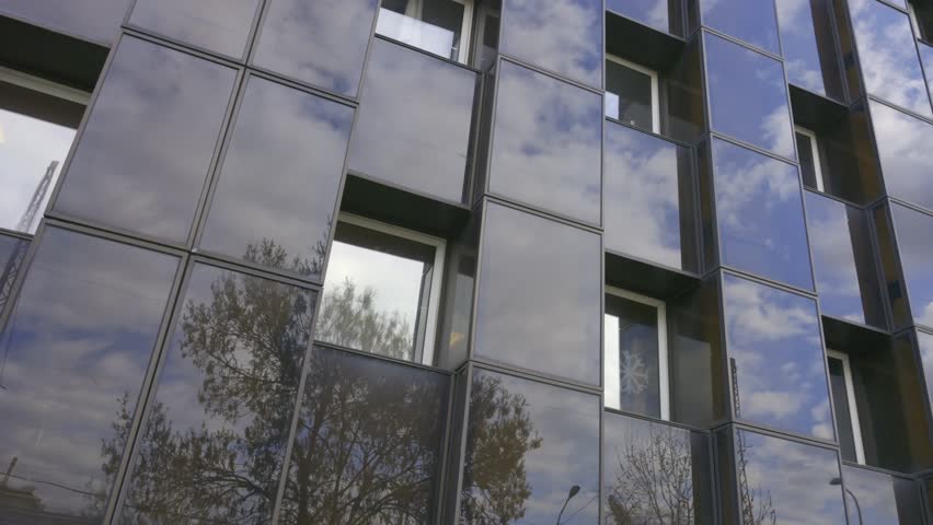 A low-angle close-up video of a modern building facade with glass cubic panels reflecting the cloudy blue sky and trees. The geometric architecture features deep black frames around windows.