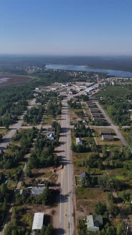 Vertical drone moving forward along Sherritt Avenue into Lynn Lake Manitoba showing abandoned houses, commercial buildings, forest, and a lake in the background on a sunny summer day.