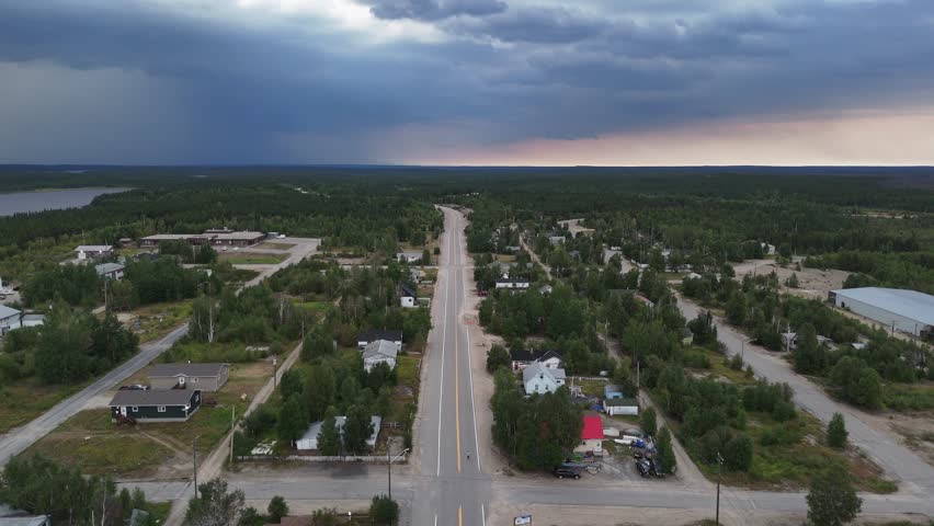 Drone flyover along a highway leaving Lynn Lake Manitoba showing residential buildings, some abandoned, with trees and cloudy skies before rain in a northern town.