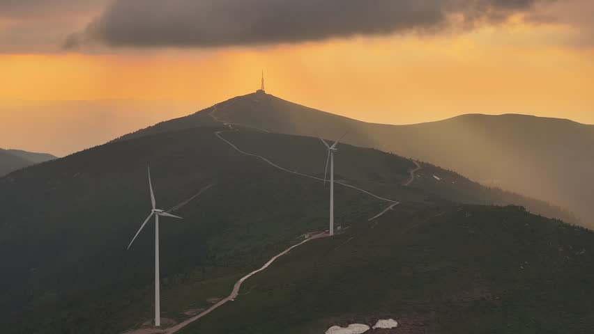 Wind turbines spin on a grassy mountain ridge against a dramatic sunset sky with rays of light