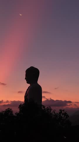 Standing Buddha statue at sunset at Tong Lam Son Temple in Nha Trang, Vietnam. Warm evening light illuminates the statue against a colorful sky creating a peaceful spiritual scene.