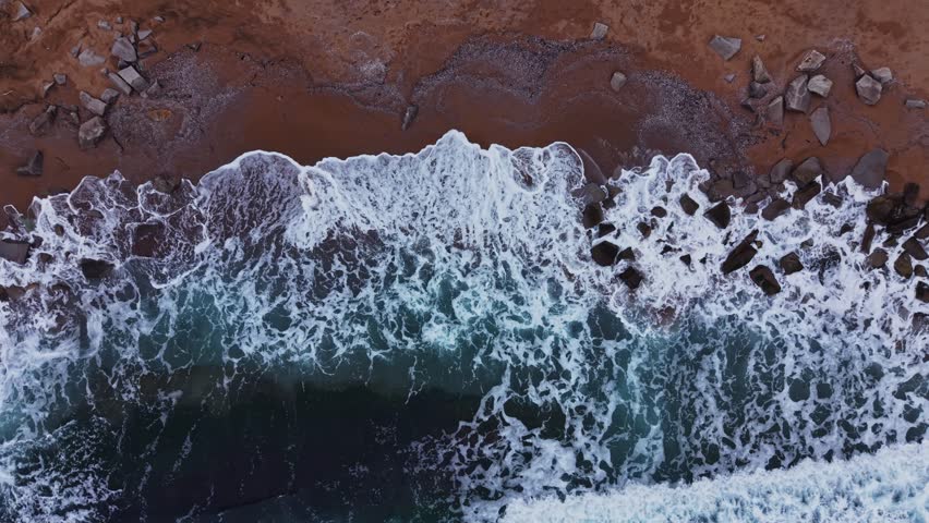 Waves crash onto a rocky shore with foam and water splashing. The coastline is visible, showing a mix of land and sea. Light reflects off the water