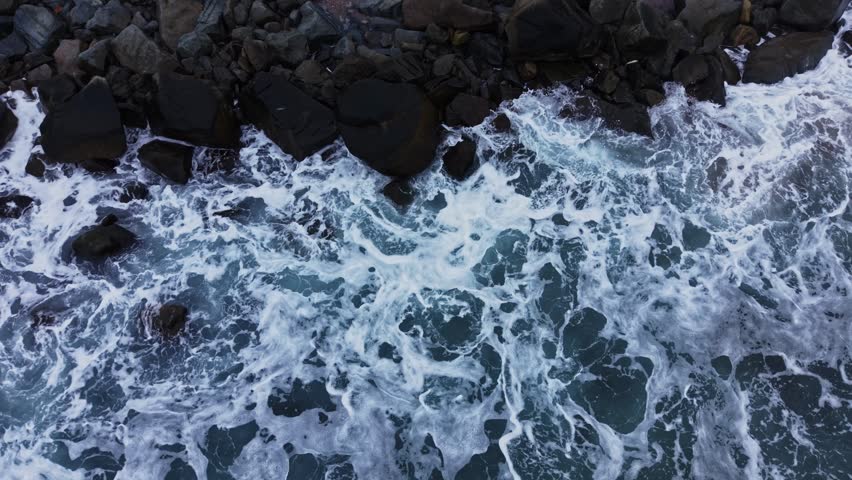 Waves roll onto a rocky shore while the sun is high. The water meets the stones, creating foamy whitecaps that contrast with the dark rocks and sandy beach below.