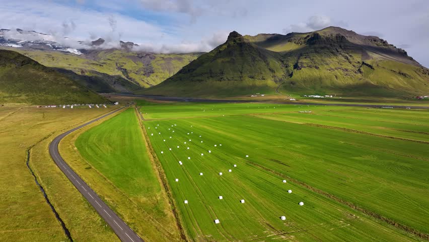 Aerial view of green farmland near Eyjafjöll in South Iceland with hay bales scattered across fields and dramatic mountains with snow patches rising in background. Wide shot.