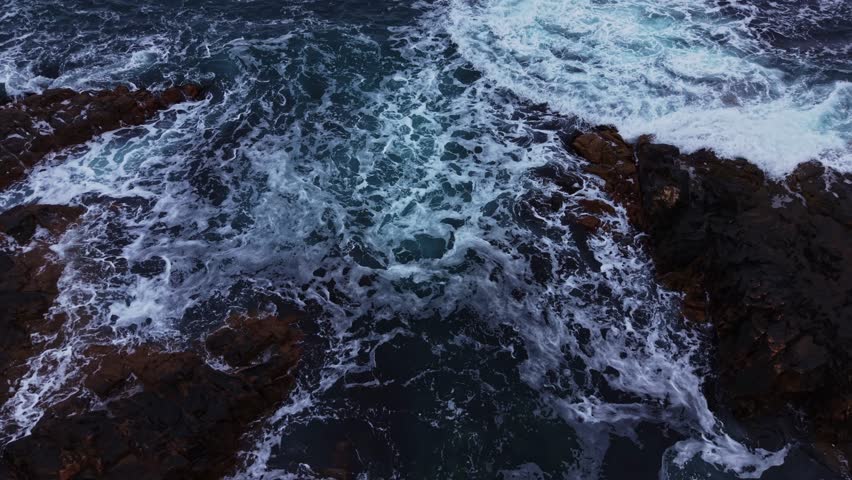 Waves move rapidly over rocks at the shoreline. Water splashes and churns as it hits the surface. The scene captures the natural motion of the ocean during daylight.