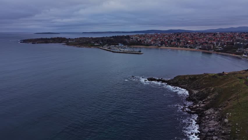 The scene shows a harbor with boats along the water. A town is visible on the shore with buildings and trees. The sky is cloudy, and the water is calm.