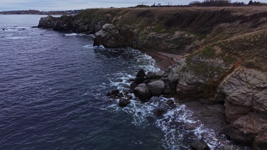 Waves crash against rocks along a rugged shoreline. The sky is cloudy, and the water reflects the grey tones of the atmosphere. A stretch of land rises in the distance.