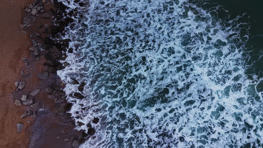 Waves crash against rocks on a beach as the sun sets. The water shows white foam and blue reflection. The shoreline is lined with sand and stones in a coastal environment.