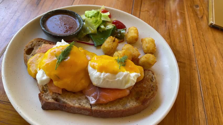 Poached eggs with hollandaise sauce over smoked salmon on sourdough toast, served with crispy potatoes and fresh salad in a café breakfast setting