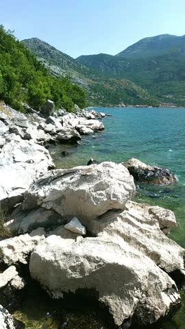 Rocky shoreline with large white stones and calm sea beneath green slopes under bright sunlight in Montenegro. Illustrates unspoiled coastal landscape, off-the-beaten-path travel, and quiet nature beyond tourist zones.